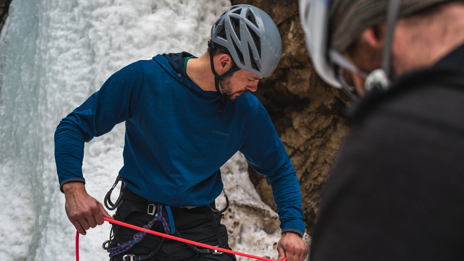 Person in VOORMI Shirt with helmet preparing for ice climbing with another person in a black shirt.